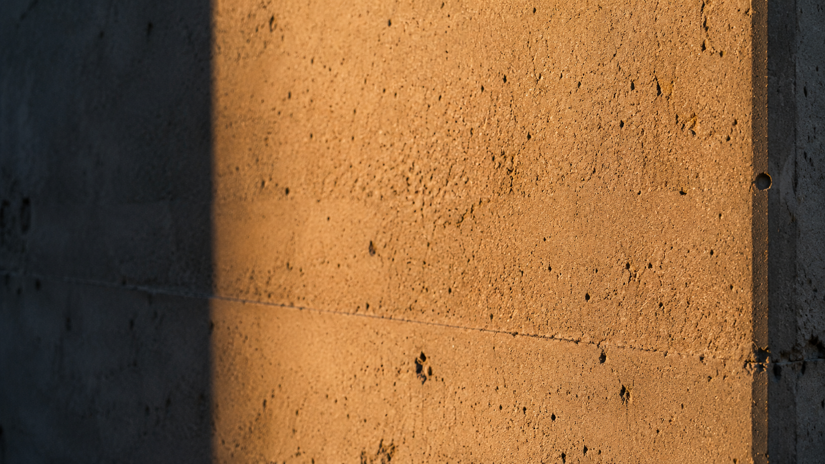 Macro photograph of raw Berlin concrete at golden hour — warm amber light raking across fine sandstone-grey surface